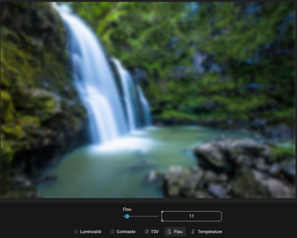 Capture d'écran d'une image (une cascade), floue, ouverte dans la partie "Réglages fins", "Flou" des modifications. dans la partie basse le paramètre pour le flou est un petit rond à tirer vers la droite ou la gauche pour modifier le contraste. Sur la droite de cette réglette, un champ avec un chiffre indiquant l'intensité du flou. En-dessous il y a 5 items, "Luminosité", "Contraste", "TSV", "Flou" et "Température".